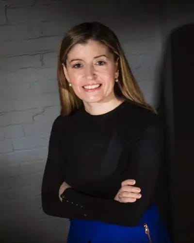 A woman with long, light brown hair smiles while standing against a brick wall.